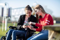 Teenage Girl In Wheelchair Looking At  Mobile Phone With Friend In Park