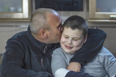 Dad and his son with autism sitting in kitchen at home. A man with short hair and bristle hugs his son and kisses him on the cheek.