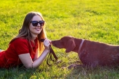 Blind woman in red clothing and sunglasses playing with the guide dog in the green grass on a sunny lawn.