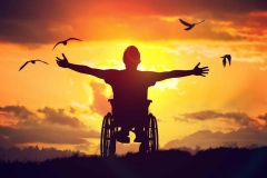 Disabled man is sitting on a wheelchair and stretching his hands at the sunset before him.