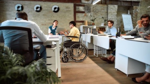 Young male worker in wheelchair talking to female colleague while presenting ideas using laptop