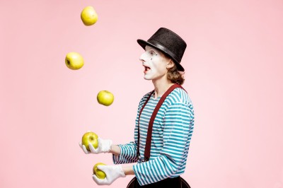 A  clown in a black and white stripy jumper and wearing a hat is juggling apples